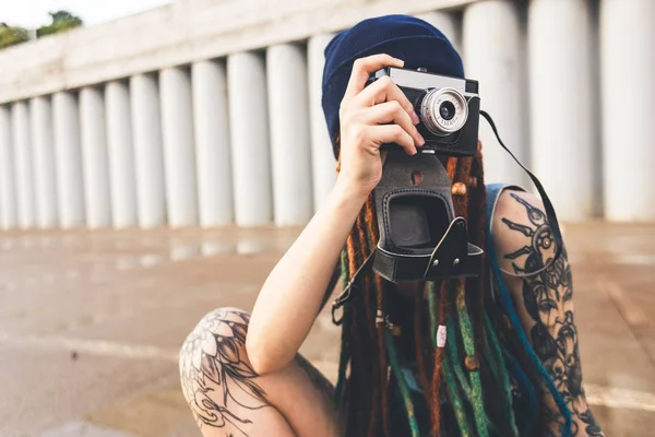 young girl with tattoos and dreadlocks in a blue hat photographs a vintage camera on the background of a concrete wall