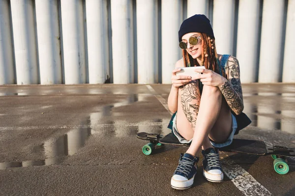 Chica joven con tatuajes y rastas en una gorra azul utiliza un teléfono inteligente contra el fondo de una pared de hormigón —  Fotos de Stock
