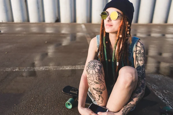 Young girl with tattoos and dreadlocks in a blue cap sits on a longboard against the background of concrete structure — Stock Photo, Image