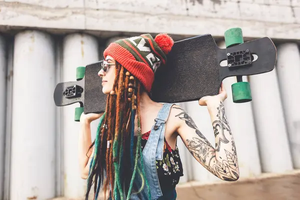 Young girl in sun glasses and a cap stands with a longboard against the background of concrete structure — Stock Photo, Image