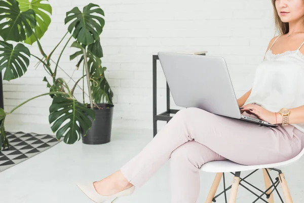 Portret van een jonge blonde meisje zittend op een stoel tegen een witte muur en werken op een laptop — Stockfoto