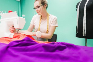 Dikiş atölyesi. Terzi iş başında. Dikiş makinesi ile renkli bir arka plan üzerine çalışan genç kadın