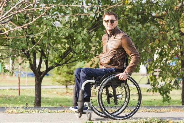 Ένας νεαρός άνδρας σε μια αναπηρική καρέκλα βόλτες κατά μήκος του δρόμου πάρκο. — Φωτογραφία Αρχείου
