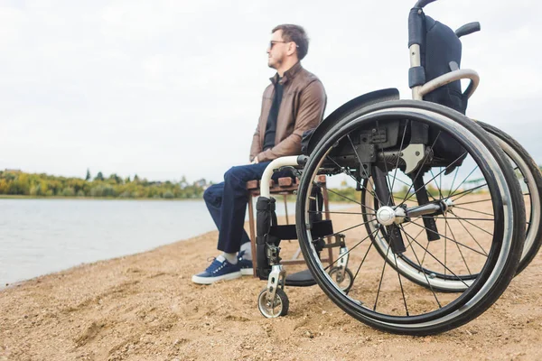 Un giovane uomo siede su una panchina vicino al lago, accanto alla sua sedia a rotelle . — Foto Stock