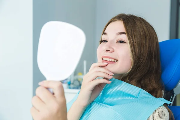 Clínica dental. Recepción, examen del paciente. Cuidado de los dientes. Chica joven sonriendo, mirando en el espejo después de un chequeo dental a su dentista — Foto de Stock