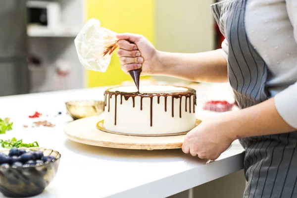 A cukrász összenyomja folyékony csokoládét egy cukrásztáskában rá egy fehér krém keksz torta egy fából készült állvány. A koncepció a házi készítésű péksütemények, főzősütemények. — Stock Fotó