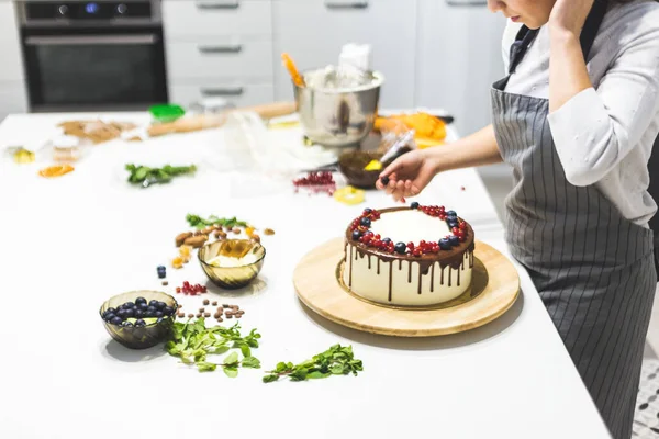 Confectioner dekorerar med bär en kak kaka med vit grädde och choklad. Kakan står på ett trä stativ på ett vitt bord. Konceptet med hembakat bak verk, matlagnings tårtor. — Stockfoto