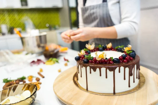 Confectioner dekorerar med bär en kak kaka med vit grädde och choklad. Kakan står på ett trä stativ på ett vitt bord. Konceptet med hembakat bak verk, matlagnings tårtor. — Stockfoto