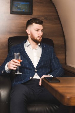 Pahalı bir takım elbiseli Genç başarılı bir işadamı elinde bir bardak şampanya ile özel bir Jet sandalyesinde oturur ve pencereden dışarı bakıyor