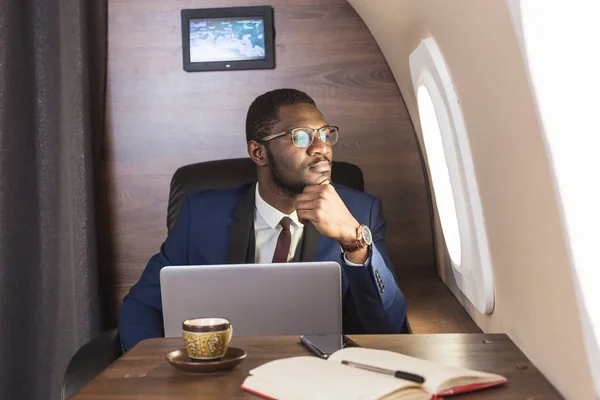 Atrakcyjny i udany African American biznesmen z okularami pracujących na laptopie siedząc na krześle jego prywatnego odrzutowca. — Zdjęcie stockowe