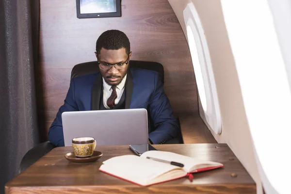 Atrakcyjny i udany African American biznesmen z okularami pracujących na laptopie siedząc na krześle jego prywatnego odrzutowca. — Zdjęcie stockowe
