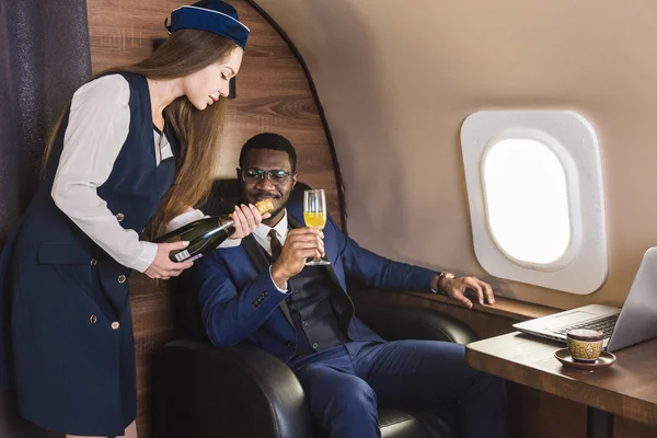 Jeune homme d'affaires afro-américain à lunettes et hôtesse de l'air montre une bouteille de vin dans la cabine d'un jet privé. Service et vol de première classe . — Photo