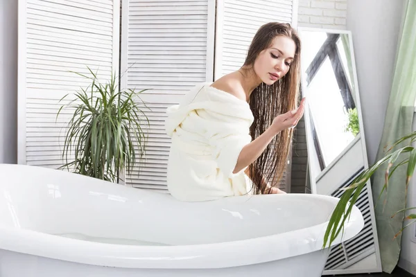 Attraente ragazza in accappatoio bianco applica la crema sulla pelle mentre si siede all'interno del bagno e facendo procedure mattutine. Cura della pelle e del corpo — Foto Stock