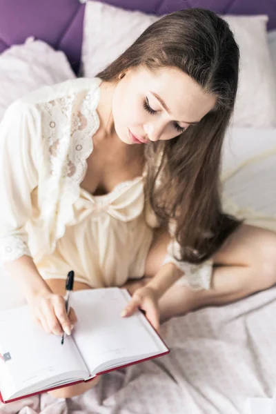 Jovem atraente em uma camisa de noite está sentado na cama e escrevendo em um caderno . — Fotografia de Stock