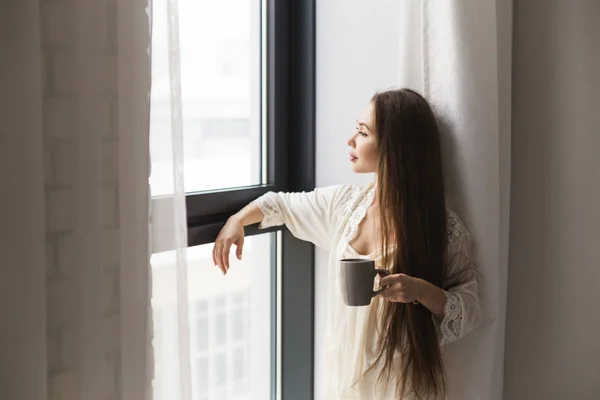 Menina atraente em roupas de noite em pé junto à janela com uma caneca de café — Fotografia de Stock