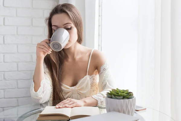 Piękna młoda dziewczyna w koszuli nocnej siedzi rano przy szklanym stole, spogląda na okno i pije kawę — Zdjęcie stockowe
