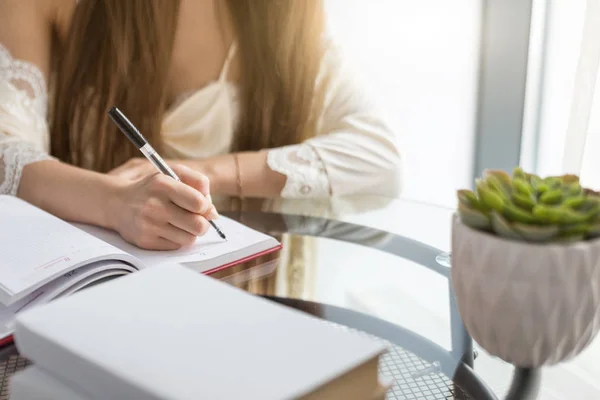 Una bella ragazza in camicia da notte è seduta al mattino a un tavolo di vetro e scrive i suoi pensieri in un quaderno. . — Foto Stock