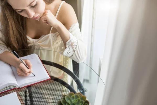 Uma menina bonita em uma camisa de noite está sentada de manhã em uma mesa de vidro e escreve seus pensamentos em um caderno . — Fotografia de Stock