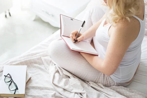 Mulher loira atraente senta-se na beira da cama em casa e escreve em um caderno — Fotografia de Stock