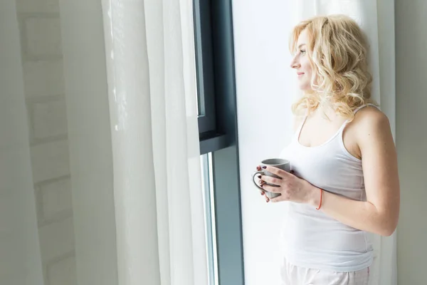 Mulher loira atraente fica perto da janela com uma xícara de café ou chá — Fotografia de Stock