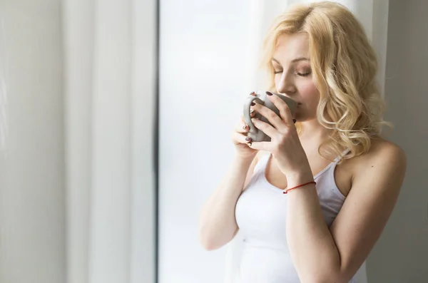 Mulher loira atraente fica perto da janela com uma xícara de café ou chá — Fotografia de Stock