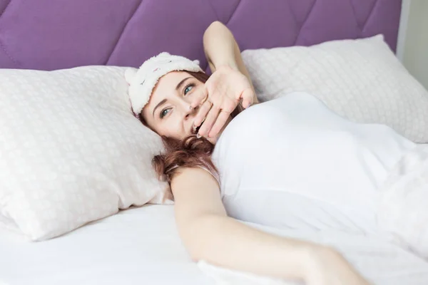 Une jolie jeune fille brune aux cheveux bruns se réveille et sirote en bâillant dans son lit dans un masque de sommeil — Photo