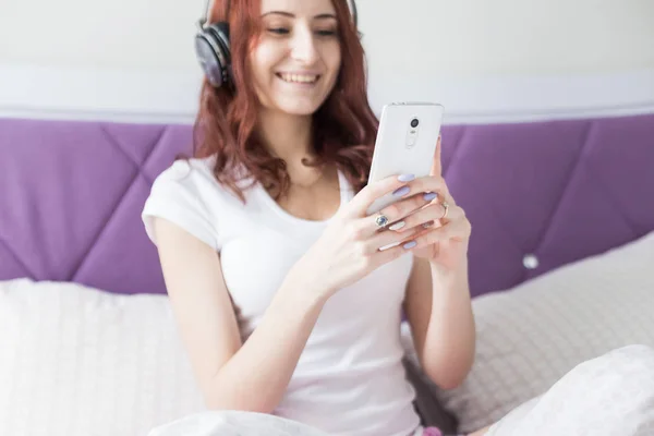 Atractiva morena joven con el pelo castaño se sienta en la mañana en su cama con auriculares y un teléfono inteligente y escucha música de buen humor — Foto de Stock