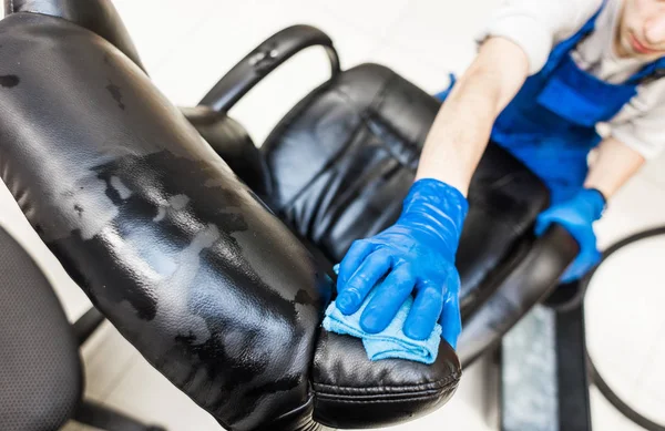 Pemuda dalam pakaian kerja dan sarung tangan karet membersihkan kursi kantor dengan peralatan profesional. — Stok Foto