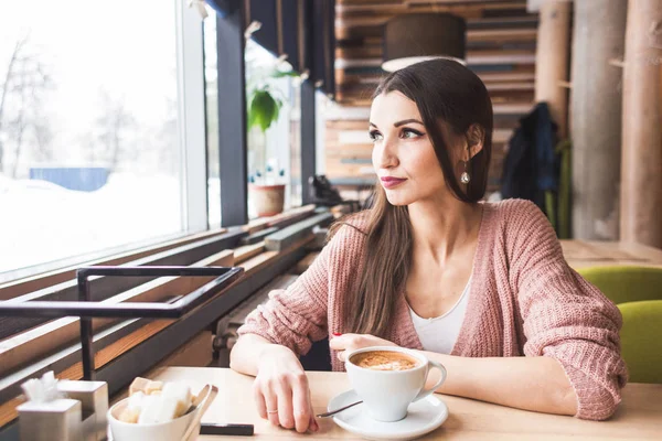 Mulher bonita se senta a uma mesa em um café com uma xícara de café e olha pela janela — Fotografia de Stock