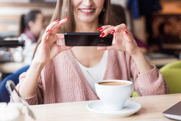 Atractiva mujer joven se sienta en una mesa en un café con una taza de café y disfruta de un ordenador portátil y teléfono inteligente —  Fotos de Stock