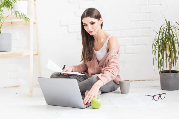 Een jonge vrolijke vrouw zit op de vloer in een licht appartement of kantoorinterieur en werkt op een laptop, freelancer girl op het werk, het concept van afstandsonderwijs studenten, papierwerk en online leren — Stockfoto