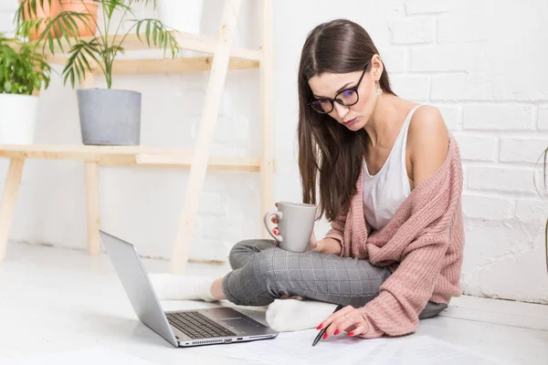 Een jonge vrolijke vrouw zit op de vloer in een licht appartement of kantoorinterieur en werkt op een laptop, freelancer girl op het werk, het concept van afstandsonderwijs studenten, papierwerk en online leren — Stockfoto