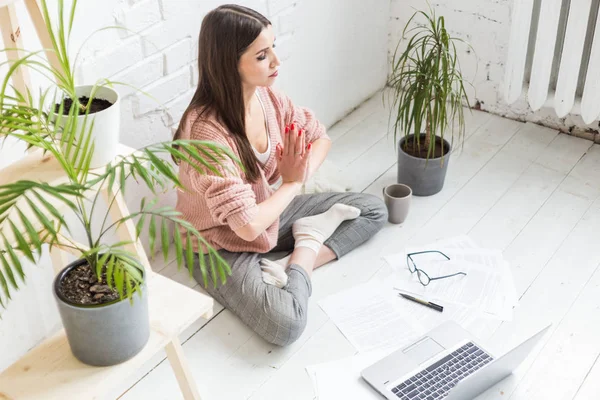 Jovem mulher feliz senta-se no chão em uma pose de ioga em um apartamento brilhante e trabalha atrás de um laptop, uma menina freelancer medita enquanto trabalha. emoção e inspiração — Fotografia de Stock