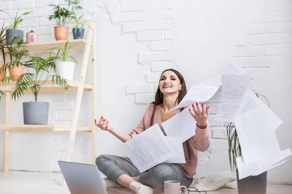 A young happy woman throws up a bunch of pieces of paper and documents sitting near the laptop, the freelance girl is happy and in a good mood. excitement and inspiration — Stock Photo, Image
