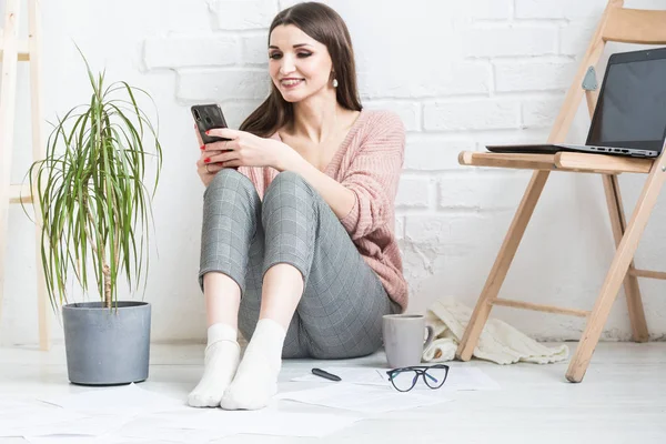 Una mujer feliz en ropa casual casera se sienta en un interior brillante y utiliza un teléfono, la chica está descansando mientras trabaja en un ordenador portátil y navegar por Internet. Acogedor espacio de trabajo independiente , — Foto de Stock