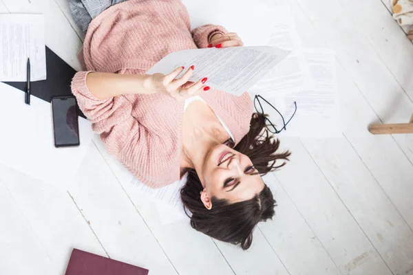 Una hermosa mujer yace en el suelo entre papeles y documentos, la chica freelancer sonríe y se relaja durante un descanso del trabajo — Foto de Stock