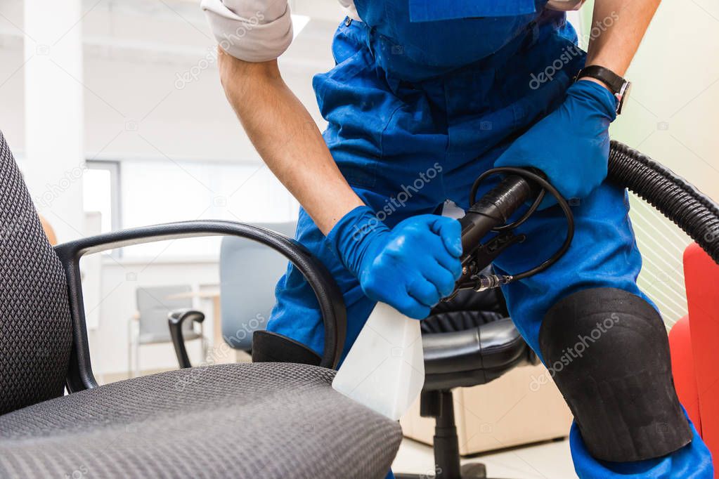 Young man in workwear and rubber gloves cleans the office chair with professional equipment.