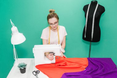 Dikiş atölyesi. Terzi iş başında. Dikiş makinesi ile renkli bir arka plan üzerine çalışan genç kadın