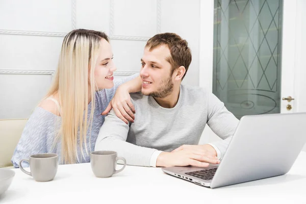 Mladí manželé sedí ráno v kuchyni a používají přenosný počítač. Online nákupní koncepce. Moderní kuchyně v pozadí. — Stock fotografie
