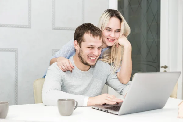 Pareja joven sentada por la mañana en la cocina y utilizando el ordenador portátil. Concepto de compras online. Cocina moderna en el fondo . — Foto de Stock