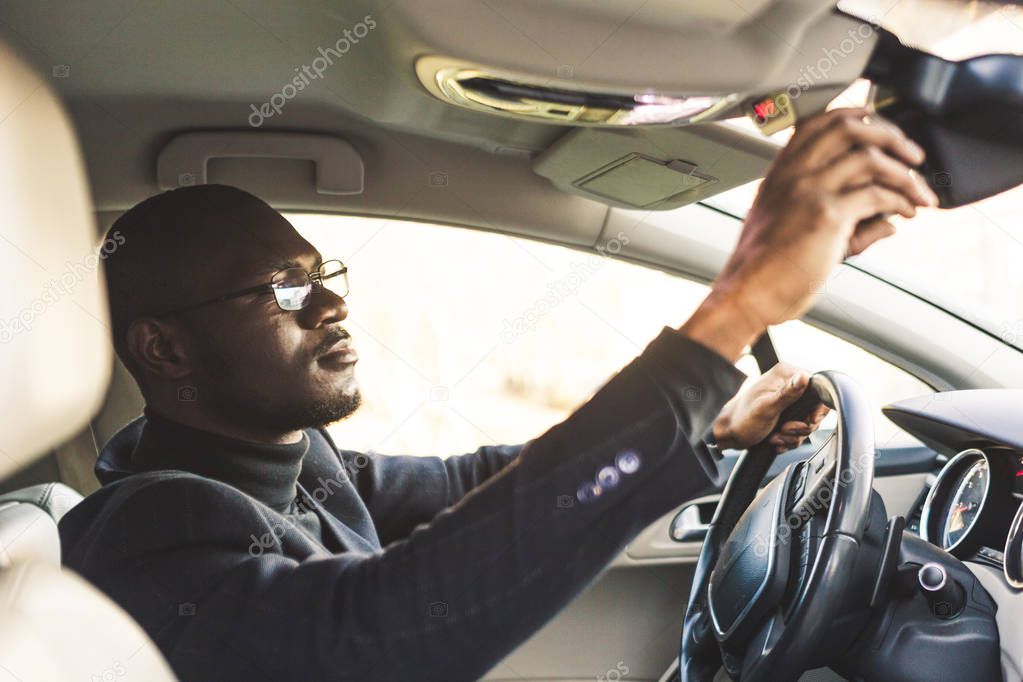 A young businessman in a suit sitting behind the wheel of a expensive car adjusts a rear view mirror.