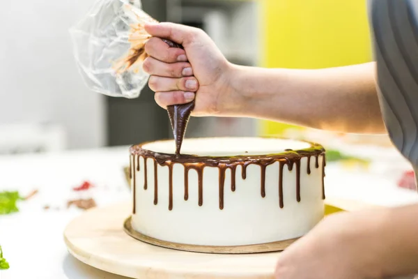 A confectioner squeezes liquid chocolate from a pastry bag onto a white cream biscuit cake on a wooden stand. The concept of homemade pastry, cooking cakes. — Stock Photo, Image