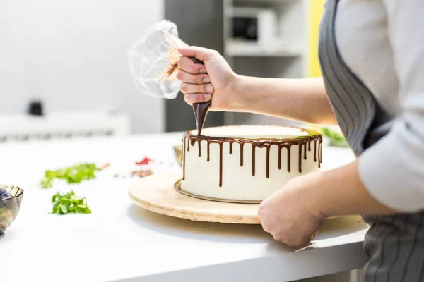 A confectioner squeezes liquid chocolate from a pastry bag onto a white cream biscuit cake on a wooden stand. The concept of homemade pastry, cooking cakes. — Stock Photo, Image