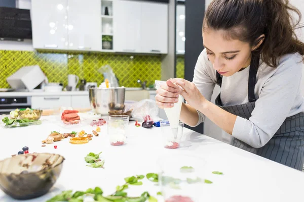 A confectioner prepares a trifle in three cups. Desserts are on the white table in the kitchen. The concept of homemade pastry, cooking cakes.