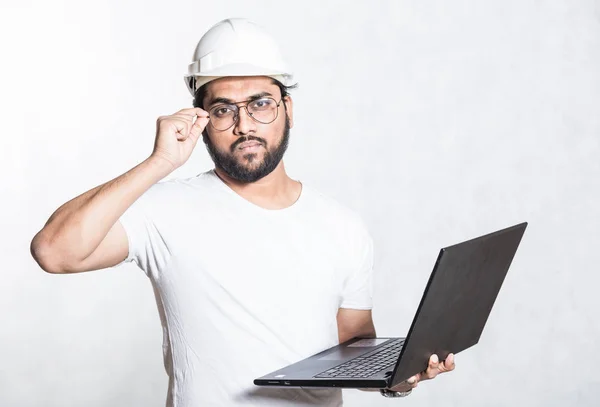 Ingeniero joven constructor en gafas y un casco de seguridad blanco, está de pie con un portátil abierto . — Foto de Stock