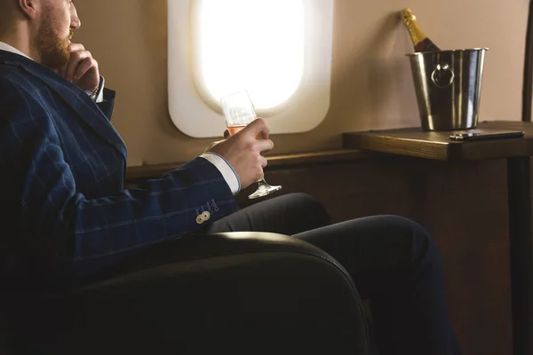 Un jeune homme d'affaires prospère en costume cher est assis sur la chaise d'un jet privé avec une coupe de champagne à la main et regarde par la fenêtre — Photo