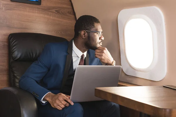 Atrakcyjny i udany African American biznesmen z okularami pracujących na laptopie siedząc na krześle jego prywatnego odrzutowca. — Zdjęcie stockowe