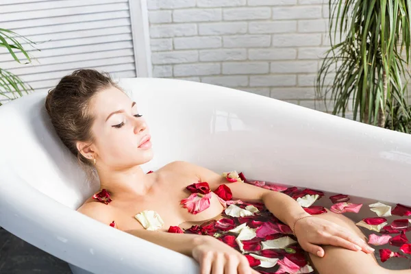 Een jong aantrekkelijk meisje neemt een badkamer met bloemblaadjes en ontspant tegen de achtergrond van een prachtig licht interieur. Spa-behandelingen voor schoonheid en gezondheid met huidverzorging — Stockfoto