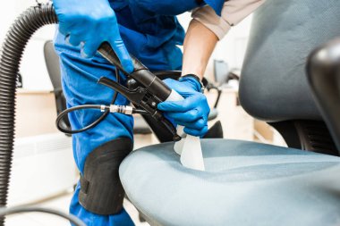 İş elbiseli ve lastik eldivenli genç adam ofis sandalyesini profesyonel ekipmanlarla temizliyor..