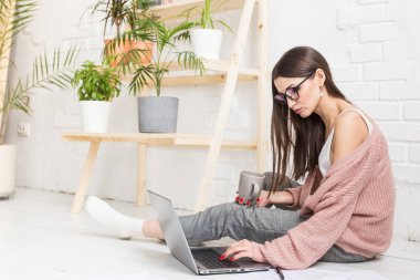 Genç ve mutlu bir kadın parlak bir apartman dairesinde ya da ofis içinde yerde oturuyor ve bir dizüstü bilgisayarda çalışıyor, iş yerinde serbest çalışan bir kız, uzaktan öğrenme öğrencileri, evrak işleri ve çevrimiçi öğrenim kavramı